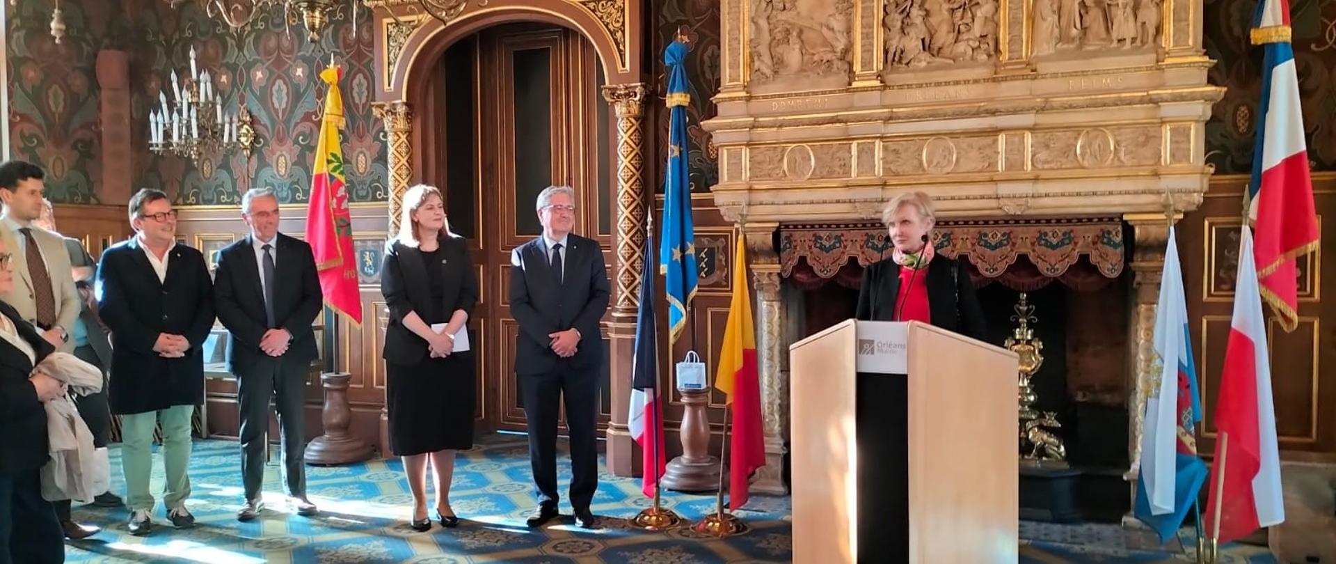 Uczestnicy spotkania słuchają na stojąco przemówienia.