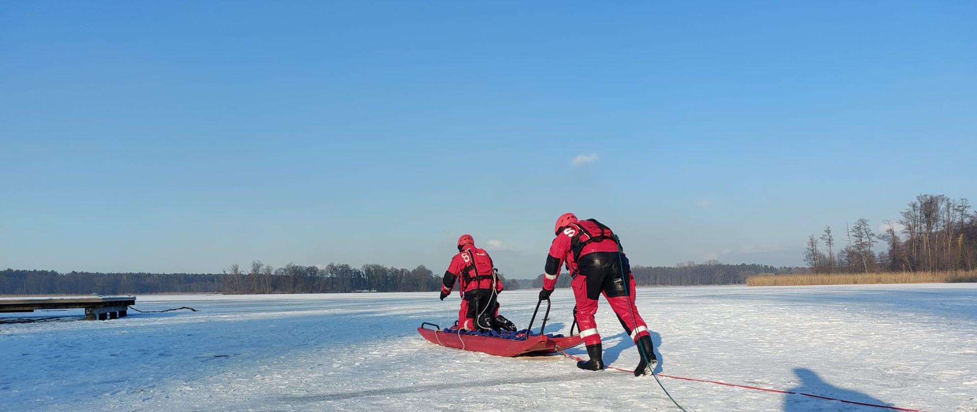 Ratownicy ćwiczą ewakuację osoby z przerębla. Na środku zdjęcia znajduje się otwór w lodzie wypełniony wodą. Dwóch ratowników w czerwonych kombinezonach leży na desce lodowej i pomaga osobie znajdującej się w wodzie. Deska jest zabezpieczona liną. W oddali, na brzegu jeziora, stoją wozy strażackie oraz strażacy obserwujący ćwiczenia. Cała scena odbywa się w zimowej scenerii, na śniegu i lodzie.
