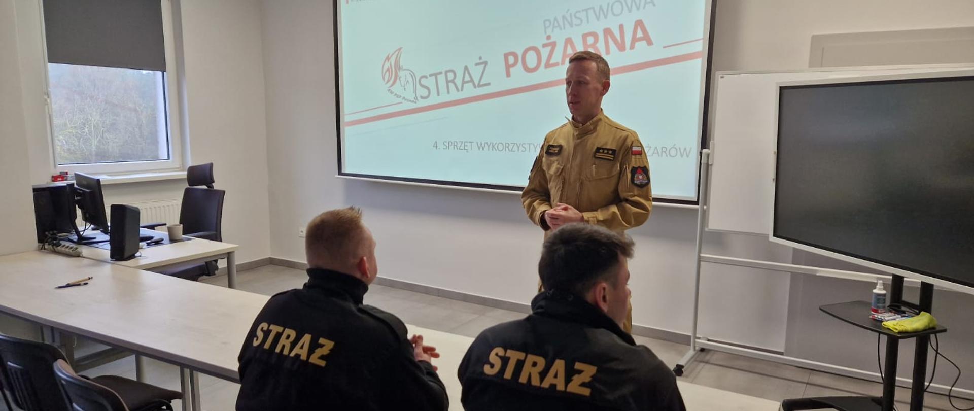 Sala wykładowa, przemawia Zastępca Wielkopolskiego Komendanta Wojewódzkiego PSP, w tle prezentacja