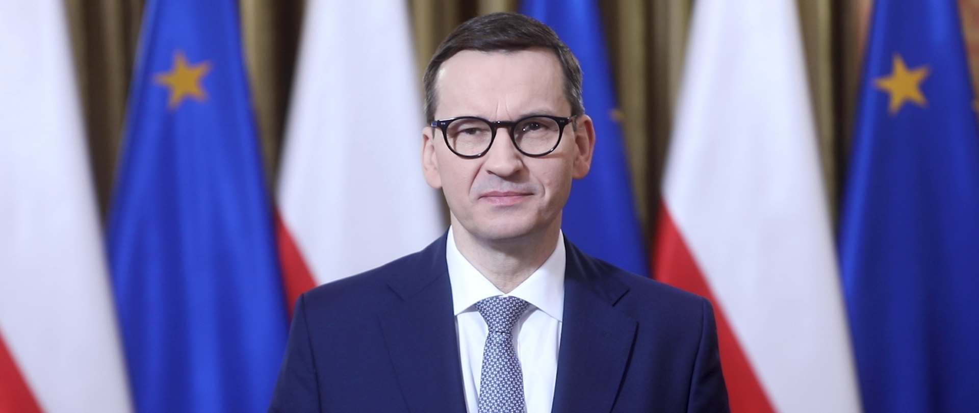 Prezes Rady Ministrów Mateusz Morawiecki na tle flag Polski i Unii Europejskiej