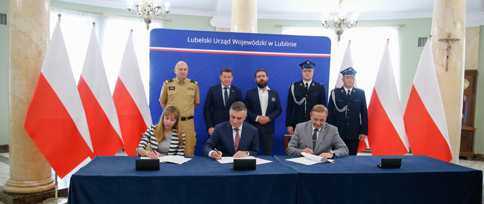 Dwóch mężczyzn i kobieta podpisują umowy. W tle biało-czerwone flagi.