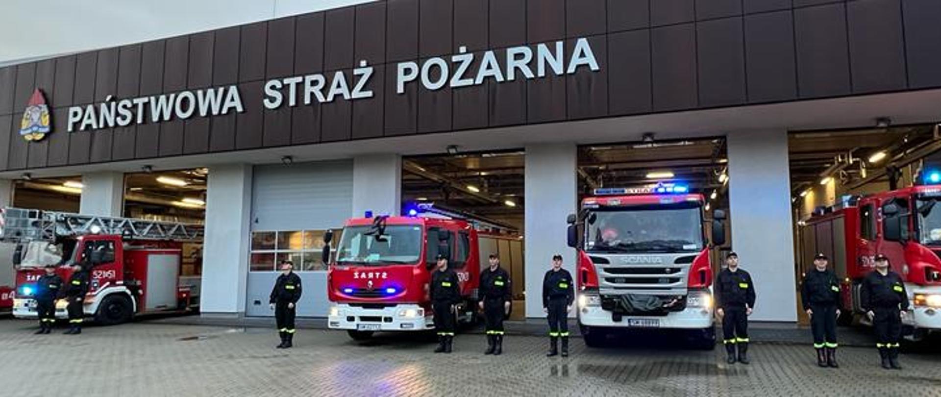 Punktualnie o godzinie 18.00 Mysłowiccy strażacy oddali hołd tragicznie zmarłemu podczas działań ratowniczych druhowi OSP Balice w tle budynek Komendy Miejskiej Państwowej Straży Pożarnej w Mysłowicach, samochody strażackie z włączonymi sygnałami i strażacy stojący przy samochodach. 