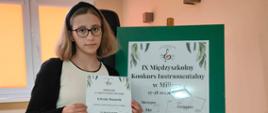 Zdjęcie przedstawia uczennicę z dyplomem na tle plakatu konkursu fletowego w Miliczu