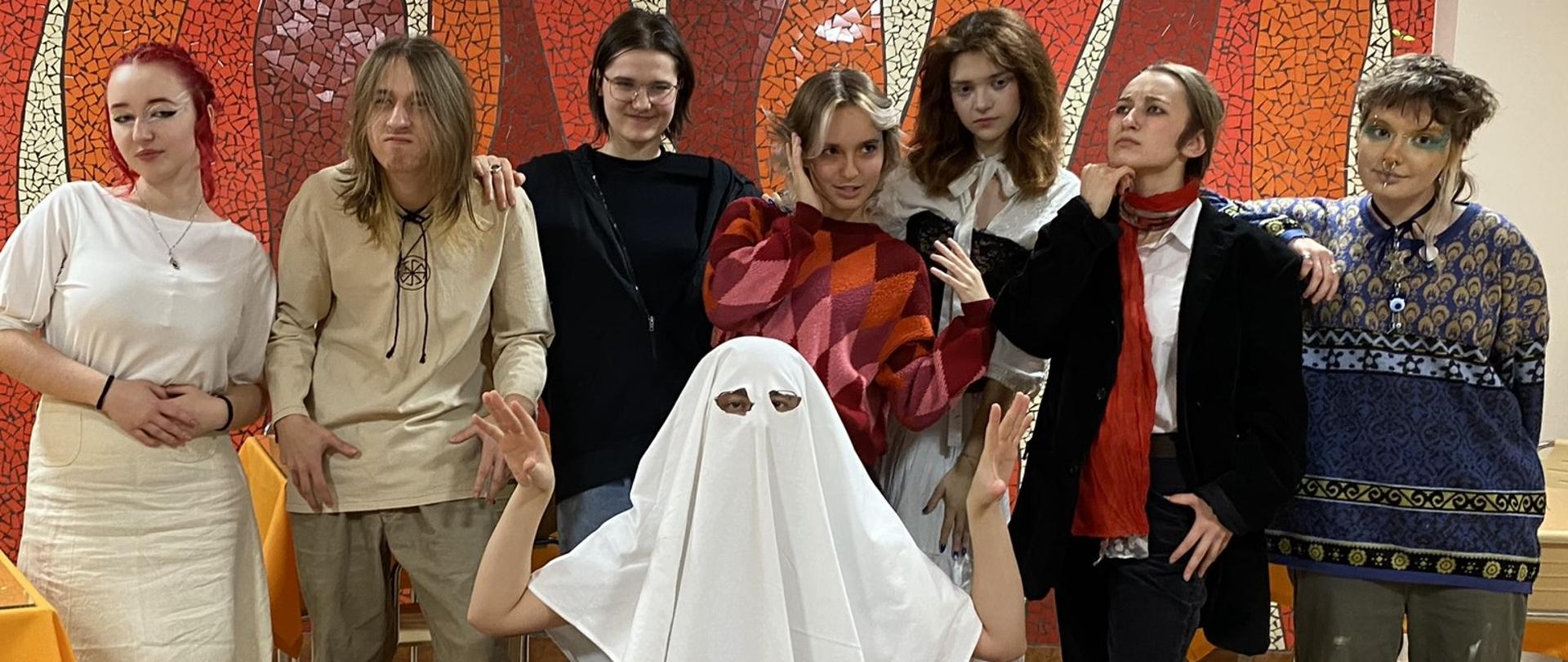 Na tle kremowo-pomarańczowo-kremowej mozaiki ściennej w stołówce pozuje ośmioosobowa grupa młodzieży przebranych za znanych artystów oraz ducha (na środku)