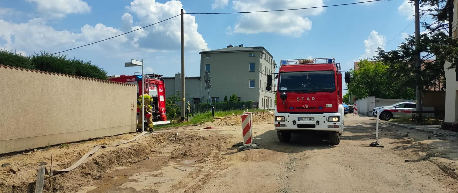 Na rozkopanej podczas remontu ulicy stoi samochód pożarniczy. Po lewej mur przyległej posesji, za którym widać tył drugiego pojazdu pożarniczego i strażaka PSP w ubraniu specjalnym. Od samochodu w kierunku patrzącego poprowadzono linie wężową do zabezpieczenia zdarzenia. W tle zabudowania. 