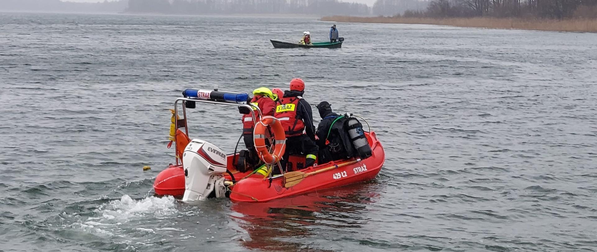 Pochmurno. Czerwona łódź ratownicza ze strażakami płynie po tafli jeziora. W tle inna łódź i drzewa