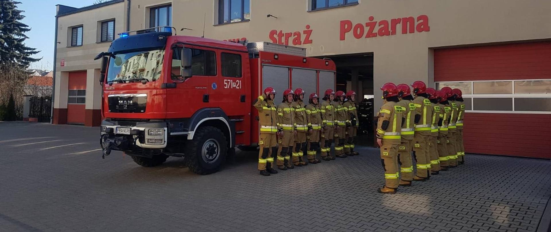 Strażacy oddali hołd kolegom z Ukrainy. Plac przed Komendą Powiatową PSP w Rawiczu. Przed budynkiem stoi wóz strażacki z włączonymi niebieskimi sygnałami błyskowymi, a obok niego strażacy z dwóch zmian służbowych. Stoją na przeciwko siebi w dwóch szeregach.