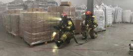 Na zdjęciu przedstawiono zadymione wnętrze hali magazynowej, gdzie składowane są palety z materiałami do produkcji plastikowych rur. Na pierwszym planie umundurowani strażacy w hełmach i aparatach oddechowych, kucając i oświetlając teren przemieszczają się z rozciągniętą linią wężową.
