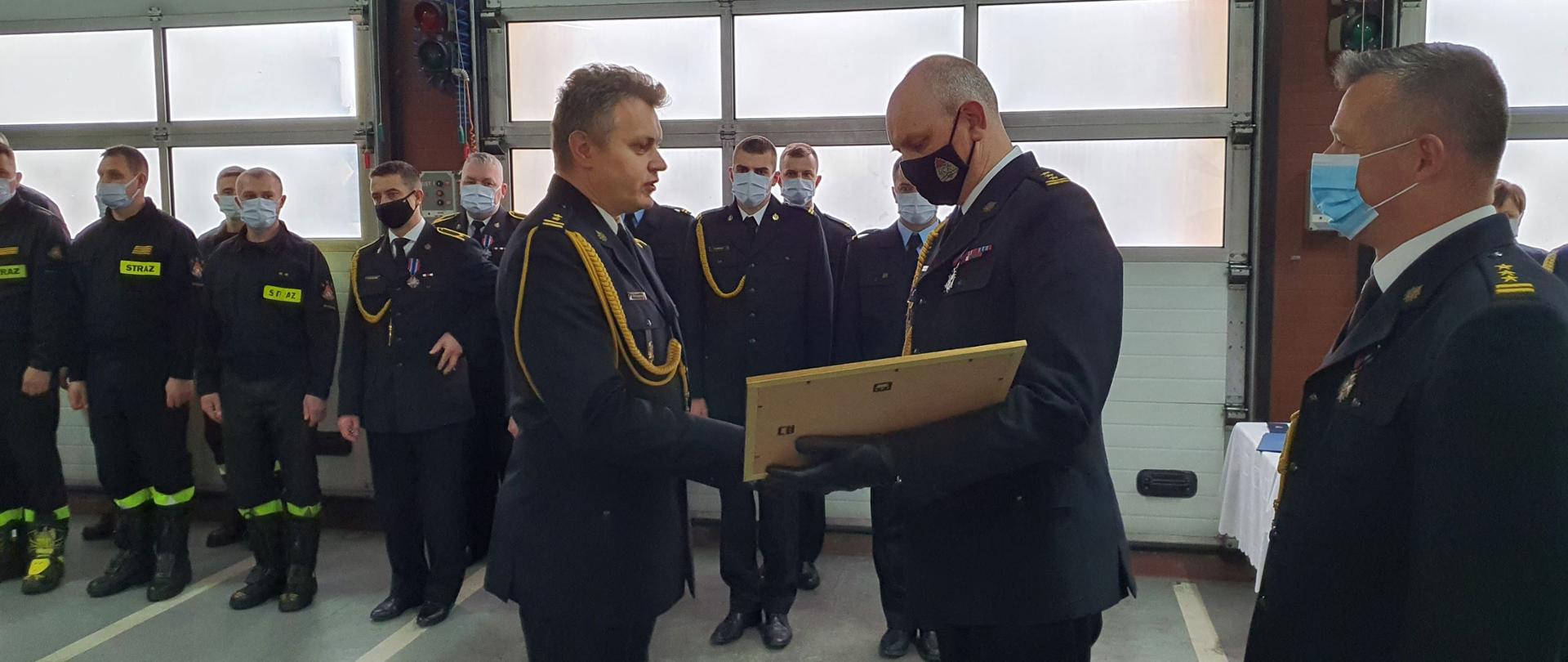 Zdjęcie przedstawia strażaków ubranych w granatowe mundury wyjściowe podczas wręczania odznaczeń i uścisków dłoni na uroczystej zbiórce w pomieszczeniu garażowym.