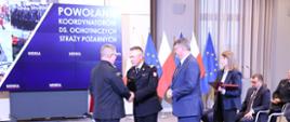 Strażak w mundurze wyjściowym odbiera gratulacje od komendanta głównego Państwowej Straży Pożarnej obok stoi sekretarz stanu w Ministerstwie Spraw Wewnętrznych i Administracji za nimi stoi kobieta z teczkami. 