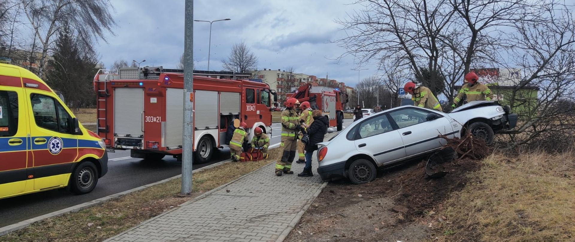 Na zdjęciu widzimy: po lewej stronie samochody służb ratowniczych kolejno: ambulans Ratownictwa Medycznego i dwa pojazdy Państwowej Straży Pożarnej. Przy samochodzie straży pożarnej dwóch strażaków udziela pomocy kierowcy uszkodzonego pojazdu. Po prawej stronie widać samochód stojący na stromym trawniku, przed maską znajduje się uszkodzone pojedyncze drzewo. Na chodniku stoi dwóch strażaków, w tym jeden rozmawia z pasażerką auta. Przy samochodzie widzimy kolejnych dwóch strażaków dokonujących oględzin zniszczonego pojazdu.