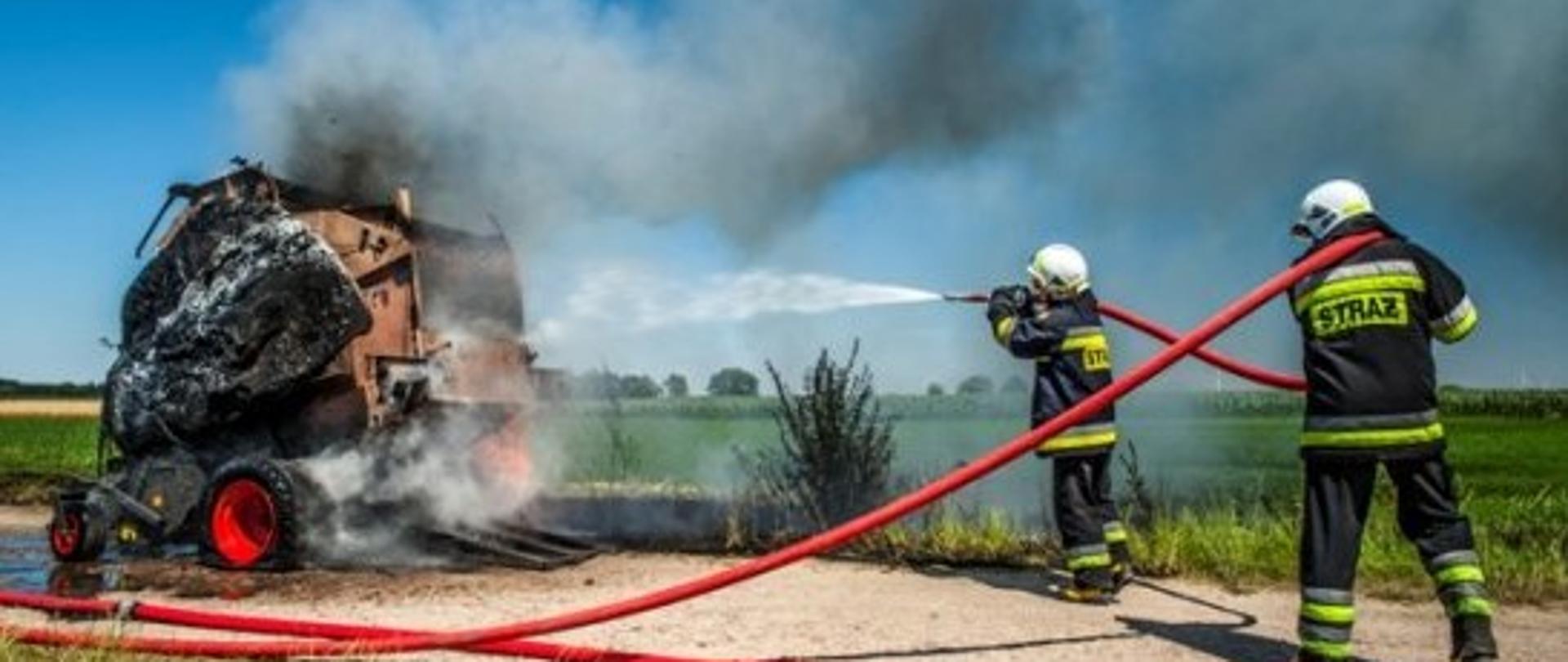 Zdjęcie archiwalne gdzie dwóch strażaków gasi prasę rolującą która zapaliła się podczas żniw. Strażacy podają jeden prąd wody.