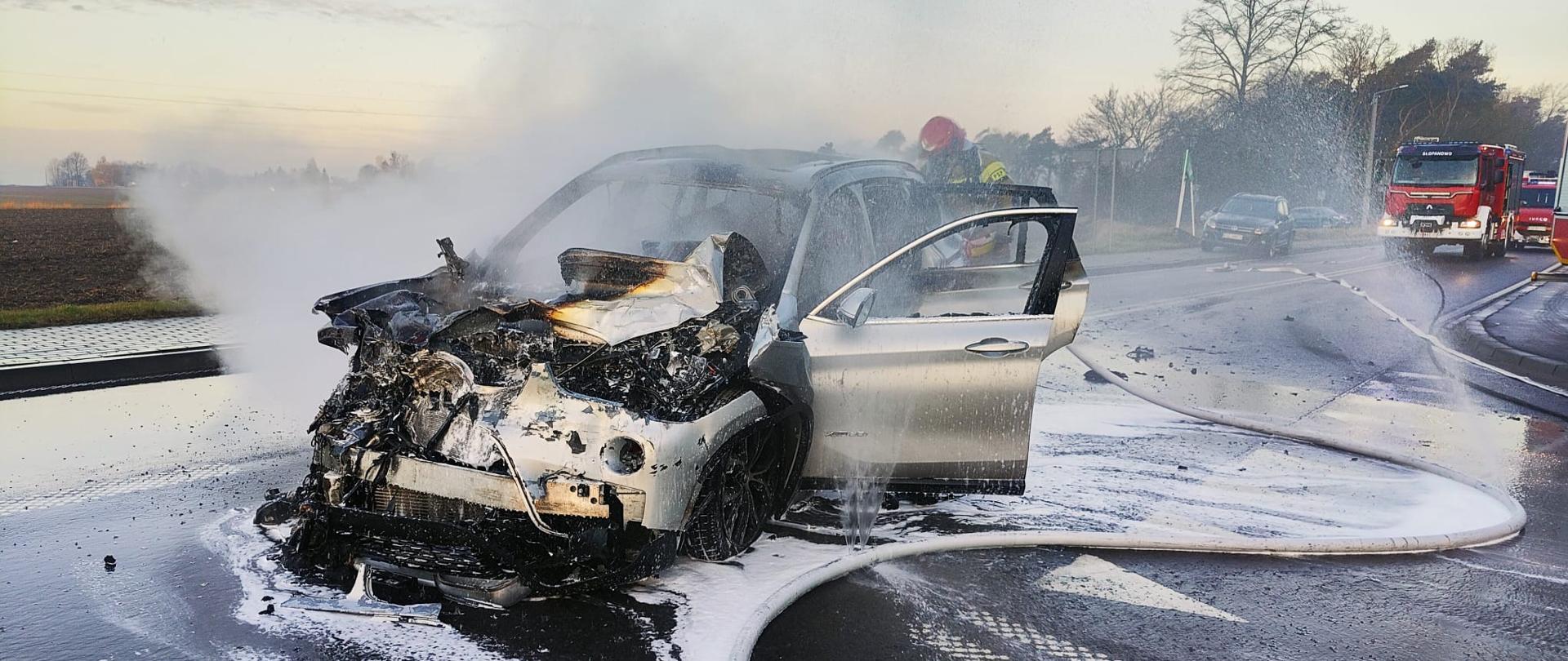 Spalony samochód na drodze, widać dym, strażak go gasi