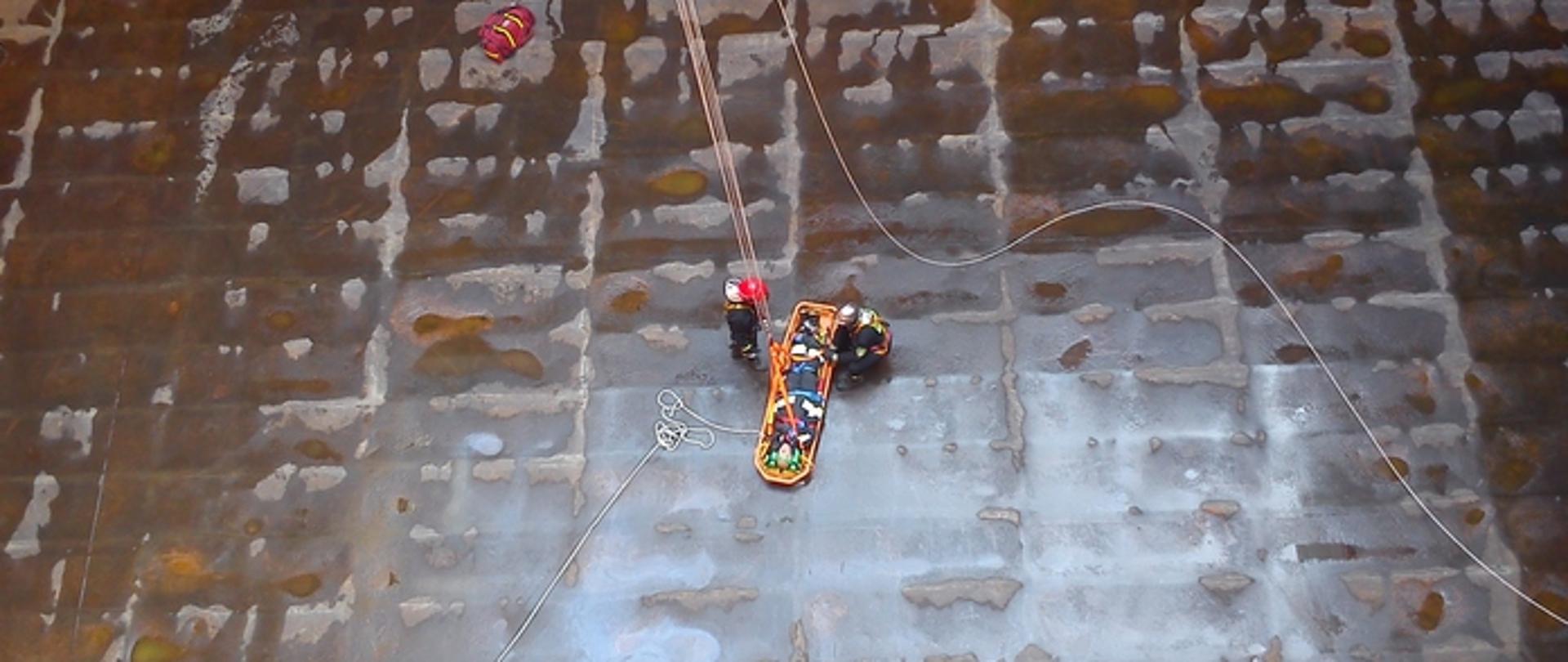 Zdjęcie przedstawia strażaków wyciągających poszkodowanego na desce z ładowni statku