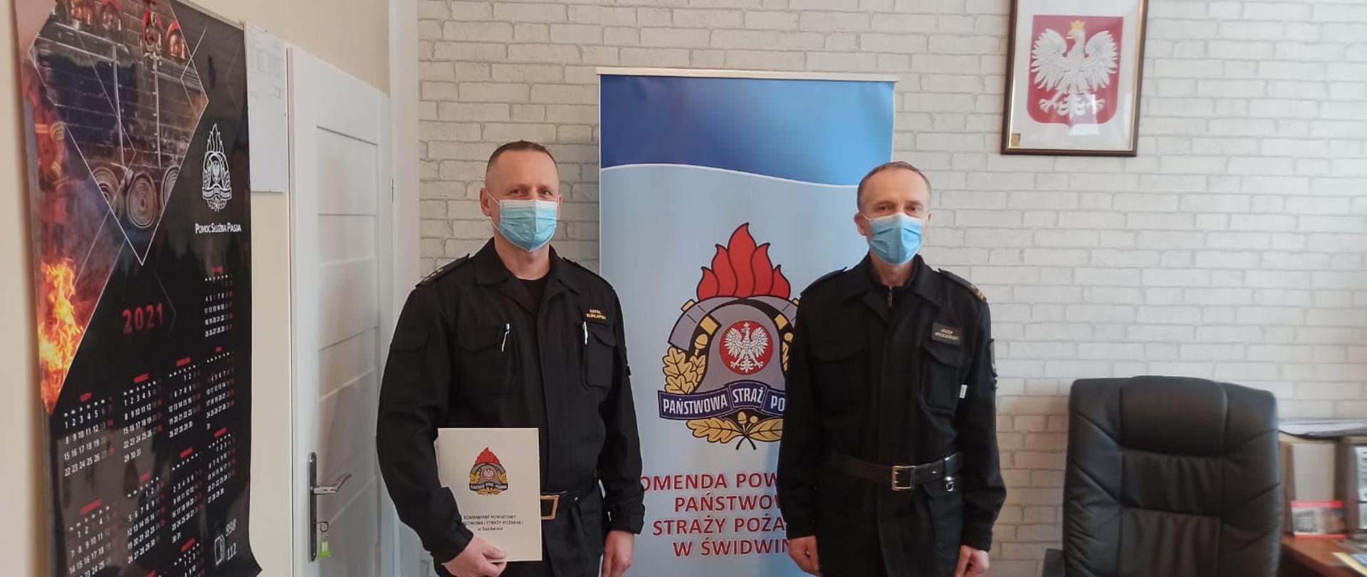 Zdjęcie przedstawia dwóch mężczyzn ubranych w czarne mundury strażackie oraz maseczki chirurgiczne, jeden z nich trzyma w rękach dokumenty. Za nimi widnieje baner z logiem PSP oraz napisem: "Komenda Powiatowa Państwowej Straży Pożarnej w Świdwinie". Za banerem widzimy ścianę z tapetą w szare cegły. Na ścianie wisi Godło Rzeczypospolitej Polskiej. Zdjęcie wykonano w gabinecie Komendanta Powiatowego PSP w Świdwinie.