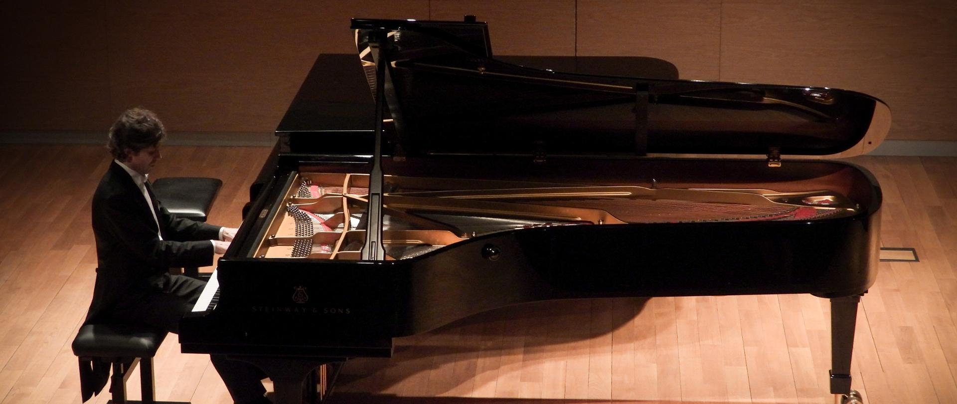 Widok z góry na pianistę grającego na czarnym fortepianie koncertowym marki Steinway & Sons. Artysta siedzi na ławie pianistycznej, a klapa instrumentu jest całkowicie otwarta, odsłaniając złociste wnętrze i struny. Scena jest wykonana z jasnego drewna, a oświetlenie skupia się punktowo na muzyku i instrumencie, tworząc nastrojowy klimat.