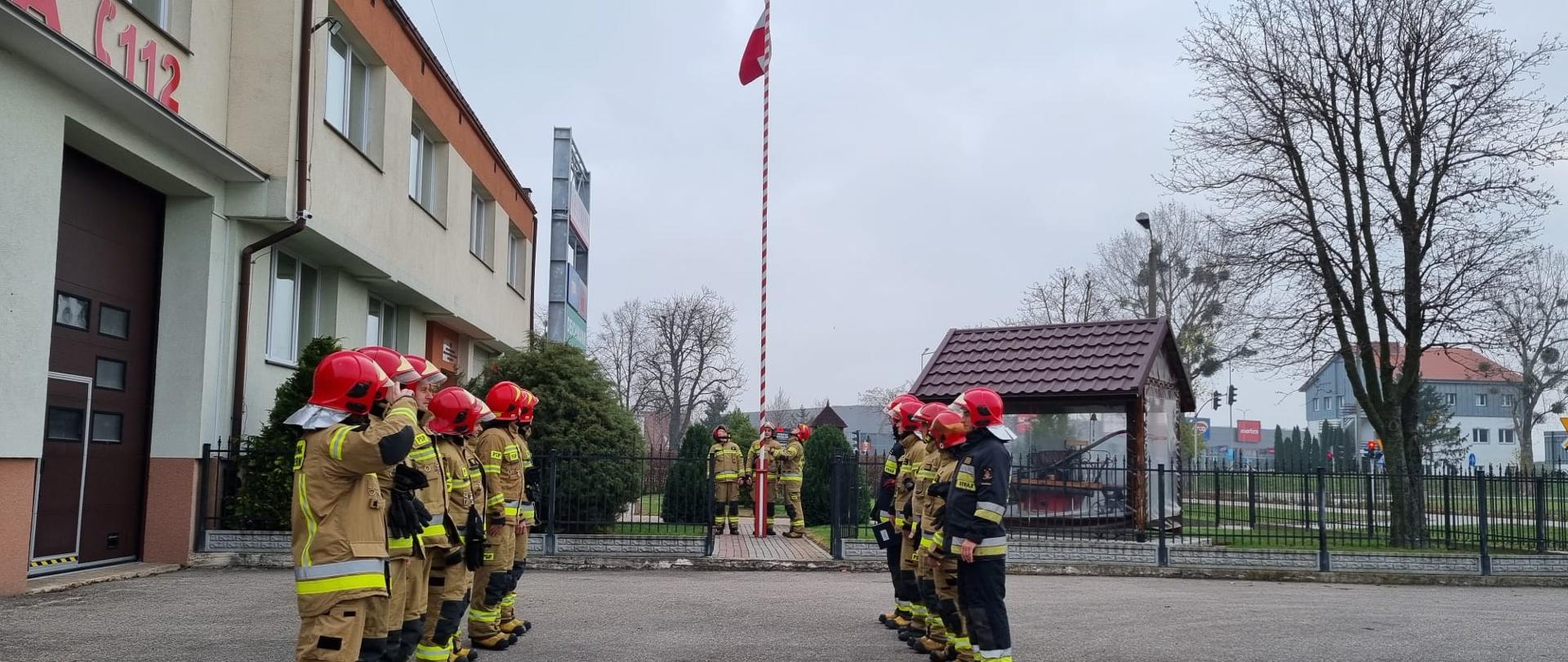 Fotografia przedstawia strażaków wciągających flagę państwową na maszt w Komendzie Powiatowej Państwowej Straży Pożarnej w Malborku.