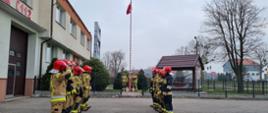 Fotografia przedstawia strażaków wciągających flagę państwową na maszt w Komendzie Powiatowej Państwowej Straży Pożarnej w Malborku.