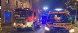 Zdjęcie wykonane w porze nocnej na miejskiej ulicy. Na ulicy samochody służb ratowniczych. Posiadają sygnalizację ostrzegawczą. W tle kamienica. W kadrze ujęta również latarnia oświetlenia ulicznego.