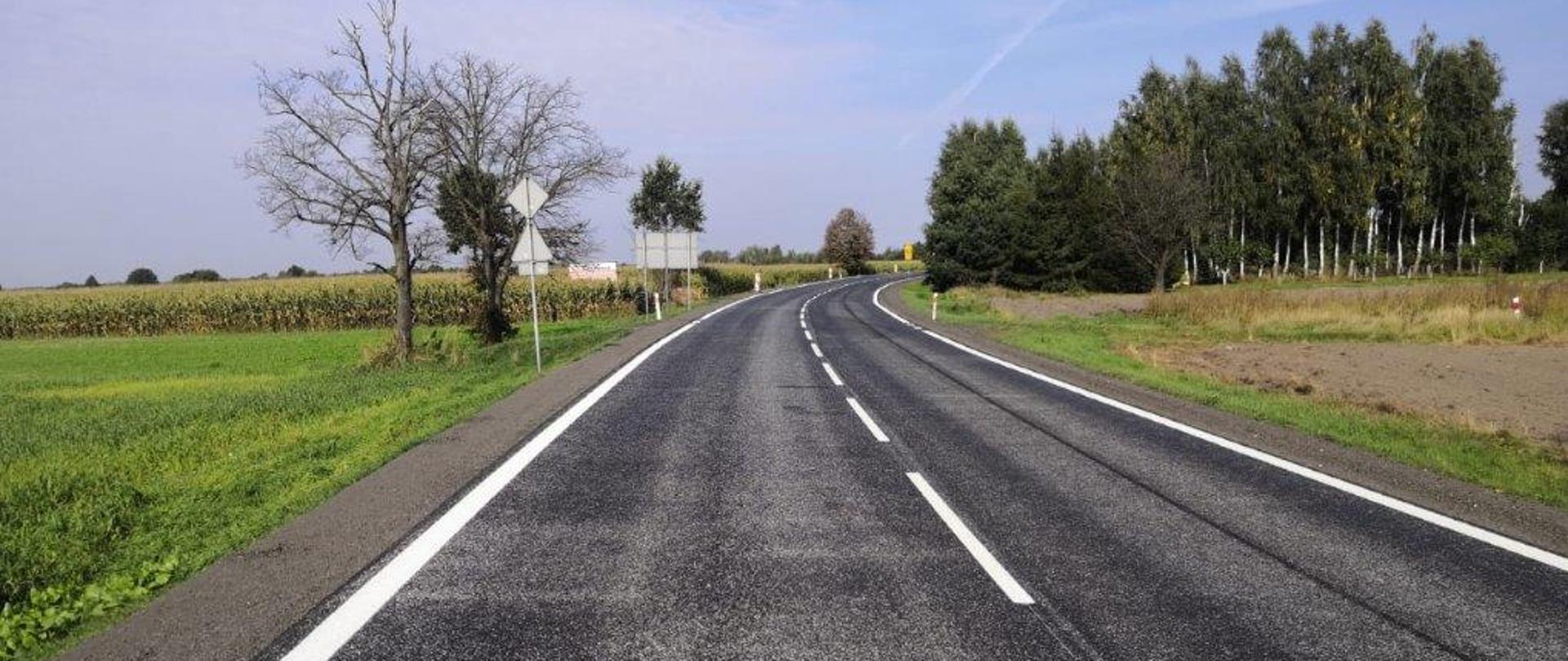 wyremontowana droga z nowym oznakowaniem poziomym, przy poboczach linia ciągła, po środku linia przerywana, w oddali po lewej pole z kukurydzą, po prawej stronie gęste zalesie.