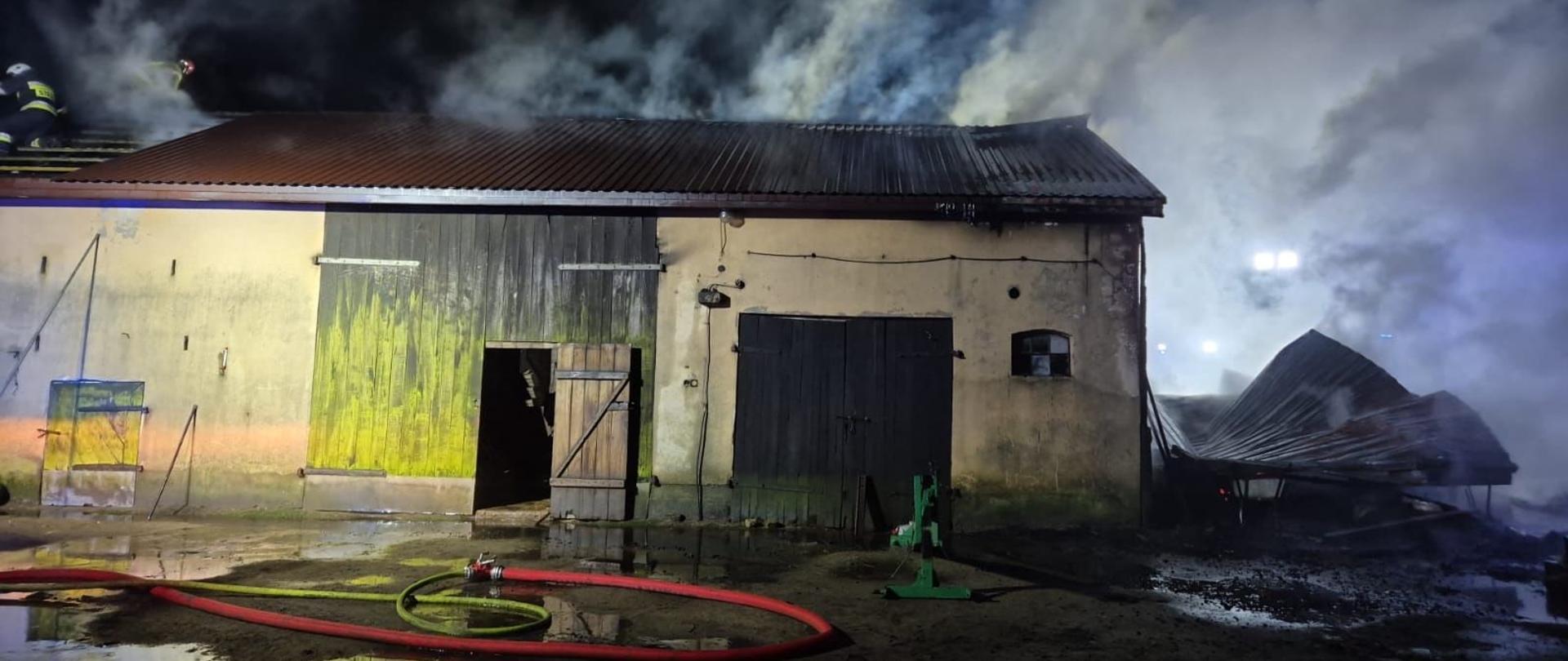 Na zdjęciu widać budynek gospodarczy, który uległ pożarowi. Scena ma miejsce nocą — teren jest oświetlony. Z dachu i wnętrza budynku unosi się gęsty dym. Część konstrukcji dachu po prawej stronie zawaliła się, a na ziemi widać mokre ślady wody i rozłożone węże strażackie. Na dachu po lewej stronie można zauważyć strażaka w akcji, pracującego przy dogaszaniu ognia. Budynek ma żółto-brązowe ściany i drewniane drzwi, na których widoczne są ślady osmolenia. Całość wskazuje na trwającą lub niedawno zakończoną akcję gaśniczą.
