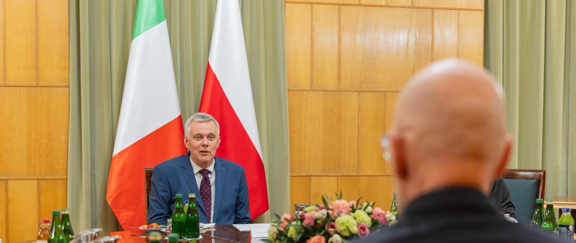 Na zdjęciu widać ministra Tomasza Siemoniaka (po lewej stronie zdjęcia) oraz ambasadora Republiki Włoskiej Lucę Franchetti Pardo (po prawej stronie zdjęcia, siedzi tyłem do obiektywu). Osoby na zdjęciu siedzą przy okrągłym, brązowym stole. Na stole m.in. zielone butelki z wodą, butelki z sokami, termosy z kawą, zastawa stołowa np. filiżanki i talerzyki. Na talerzykach ciasteczka i owoce. Na środku stołu kompozycja kwiatowa. Za ministrem stoją dwie flagi - po prawej stronie biało-czerwona flaga Polski, a po lewej stronie zielono-biało-czerwona flaga Włoch. W tle drewniane wykończenie ścian oraz dwie zielone kotary.