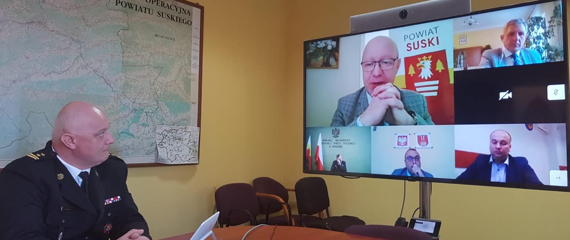 Zdjęcie przedstawia Komendanta Powiatowego PSP w Suchej Beskidzkiej. Z prawej strony znajduje się telewizor, a na min wyświetlane są osoby biorące udział w wideokonferencji. 