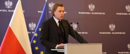 Wojewoda mazowiecki Mariusz Frankowski stoi przy mównicy, w tle ścianka, flagi polska i unijna 
