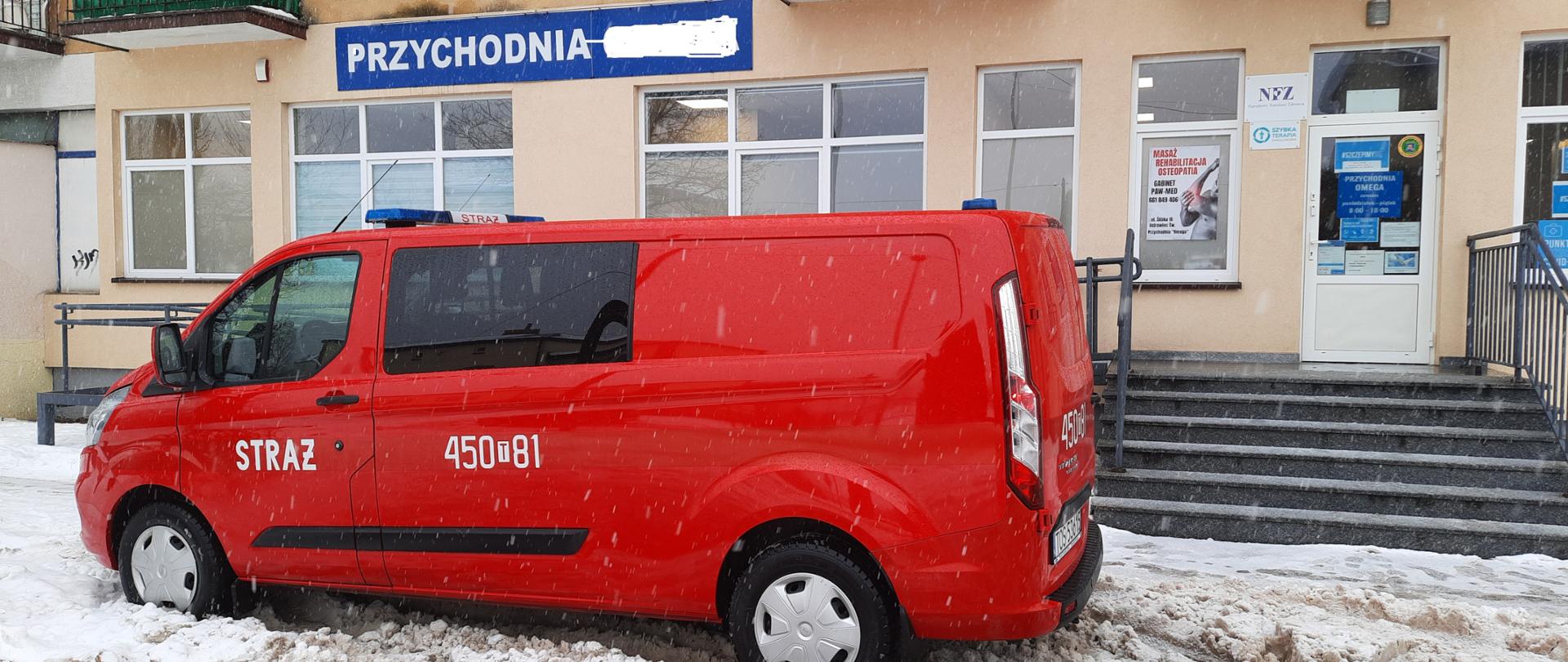 Przed przychodnią zdrowia stoi zaparkowany samochód osobowy pożarniczy. Aura zimowa. Przychodnia znajduje się na parterze w budynku mieszkalnym wielorodzinnym.