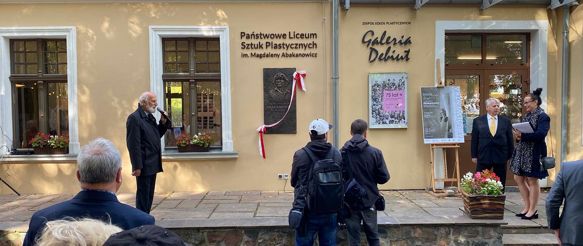 Zdjęcie przedstawia moment odsłonięcia tablicy pamiątkowej. Na tle elewacji budynku stoi dyrektor ZSP Marek Lis z mikrofonem w dłoni, po prawej stronie fotografii w odległości 5 metrów od dyrektora stoi senator Bogdan Borusewicz i nauczycielka ZSP Ewa Manuszewska. Pomiędzy postaciami znajduje się tablica pamiątkowa z płaskorzeźbą Magdaleny Abakanowicz.