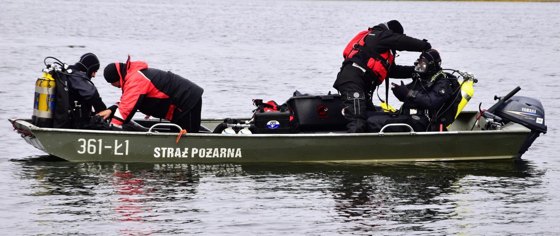 Nurkowie straży pożarnej siedzą ubrani w akwalungi na łodzi. W tle jezioro oraz lesisty brzeg.