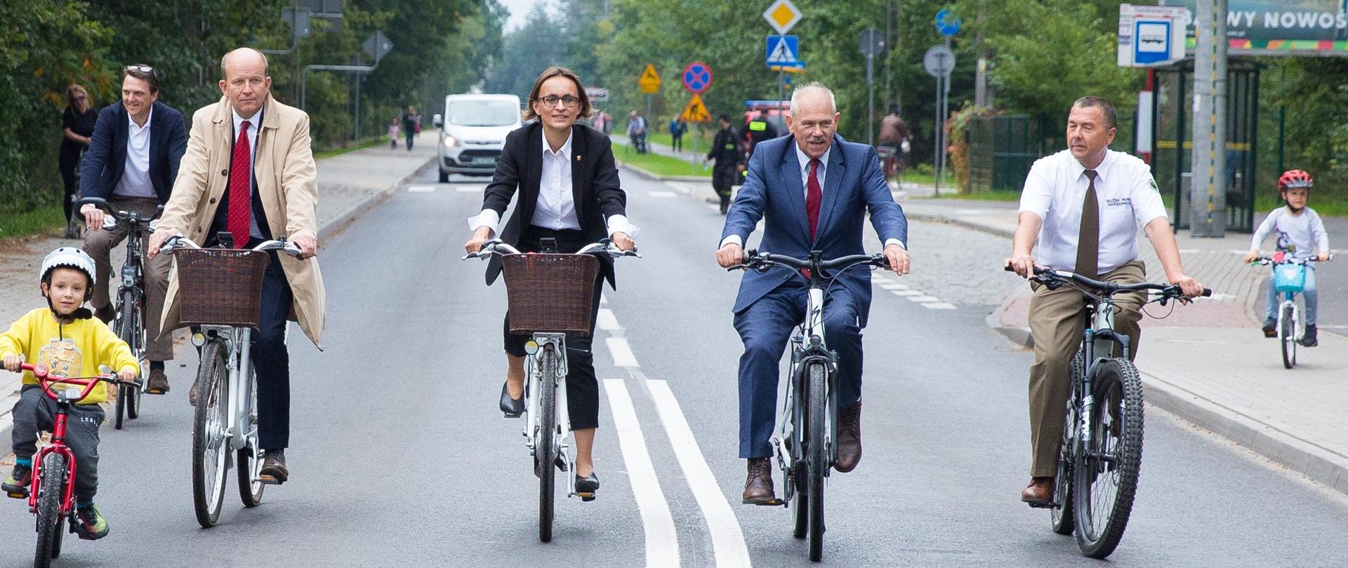 Zdjęcie przedstawia przejazd rowerowy nowo zmodernizowaną drogą powiatową. Rowerami przemieszcza się siedem osób, w tym dwójka dzieci. W tle nadjeżdżający samochód, elementy inżynierii drogowej (m.in. znaki drogowe, przystanek autobusowy). Droga przebiega przez Kampinoski Park Narodowy
