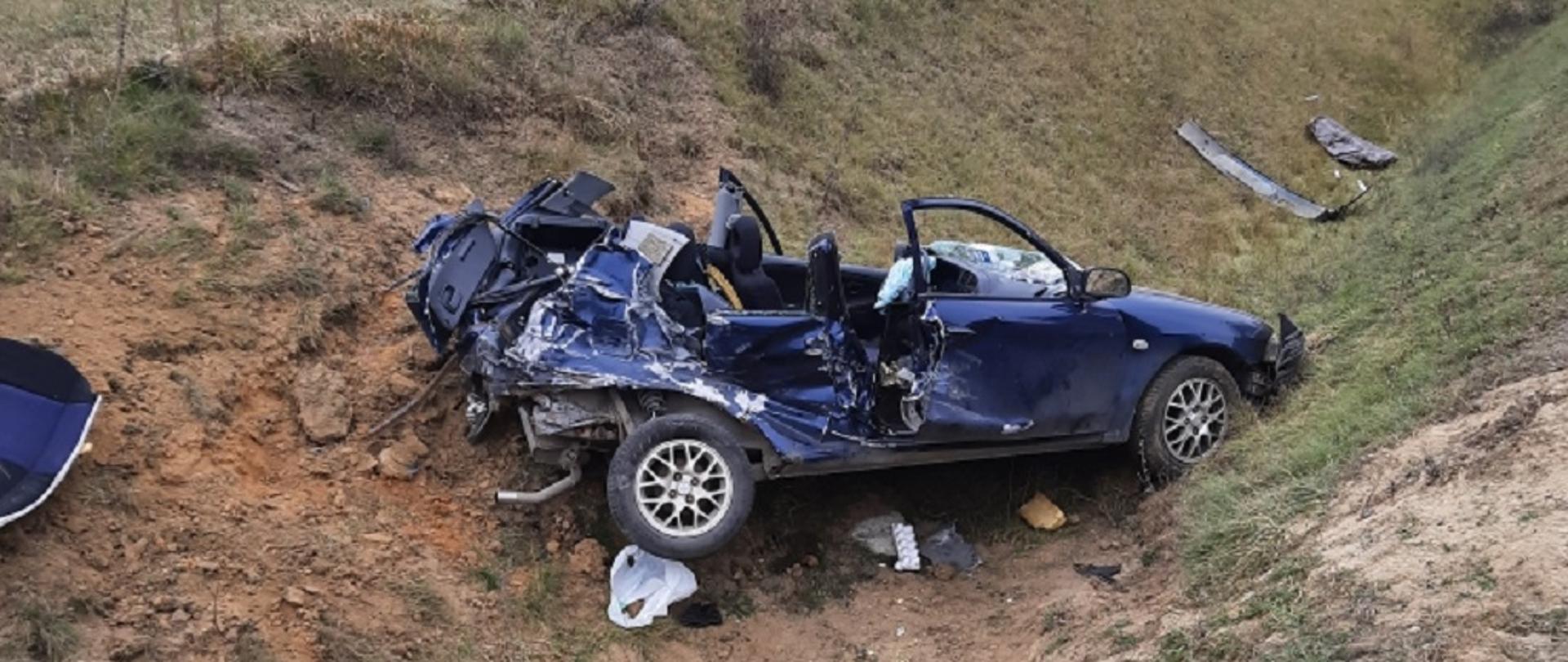 W rowie wrak niebieskiego samochodu osobowego bez dachu. W tle samochód strażacki.