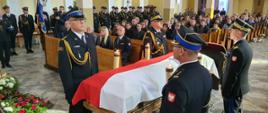 Zdjęcie przedstawia wnętrze kościoła, w którym 4 strażacy ubrani w czarne mundury galowe stoją przy trumnie. W tle widoczne ławki z siedzącymi w nich ludźmi oraz pododdział strażaków w galowych mundurach z pocztem sztandarowym
