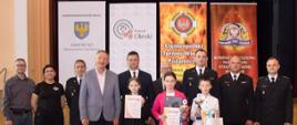 W sali w Miejskim Domu Kultury w Oleśnie są ustawieni w szeregu laureaci I kategorii wiekowej etapu powiatowego OTWP, organizatorzy i zaproszeni goście. W tle stoją 4 banery: Samorządu Województwa Opolskiego, Starostwa Powiatowego, OTWP, Komendy Powiatowej Państwowej Straży Pożarnej w Oleśnie.