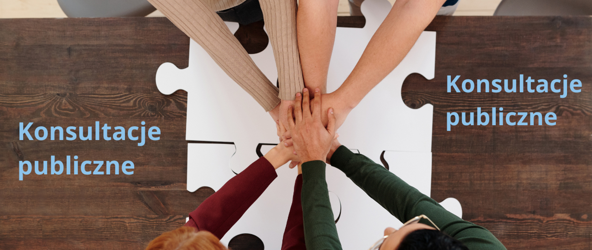 Drewniany blat stołu, na którym położone są 4 duże puzzle nad nimi połączone ręce kilku osób symbolizujące gest solidarności.