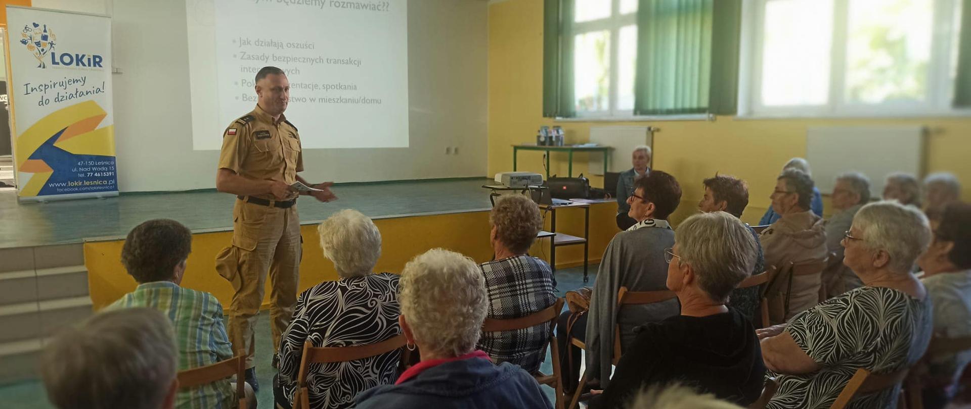 Na zdjęciu znajduje się strażak ubrany w ubranie służbowe, na krzesłach siedzą starsze osoby