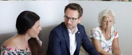 Minister rozwoju i technologii Waldemar Buda na spotkaniu z mieszkańcami osiedla przy ul. Kolejowej w Zgierzu, minister siedzi na kanapie w mieszkaniu, obok niego siedzą dwie kobiety 