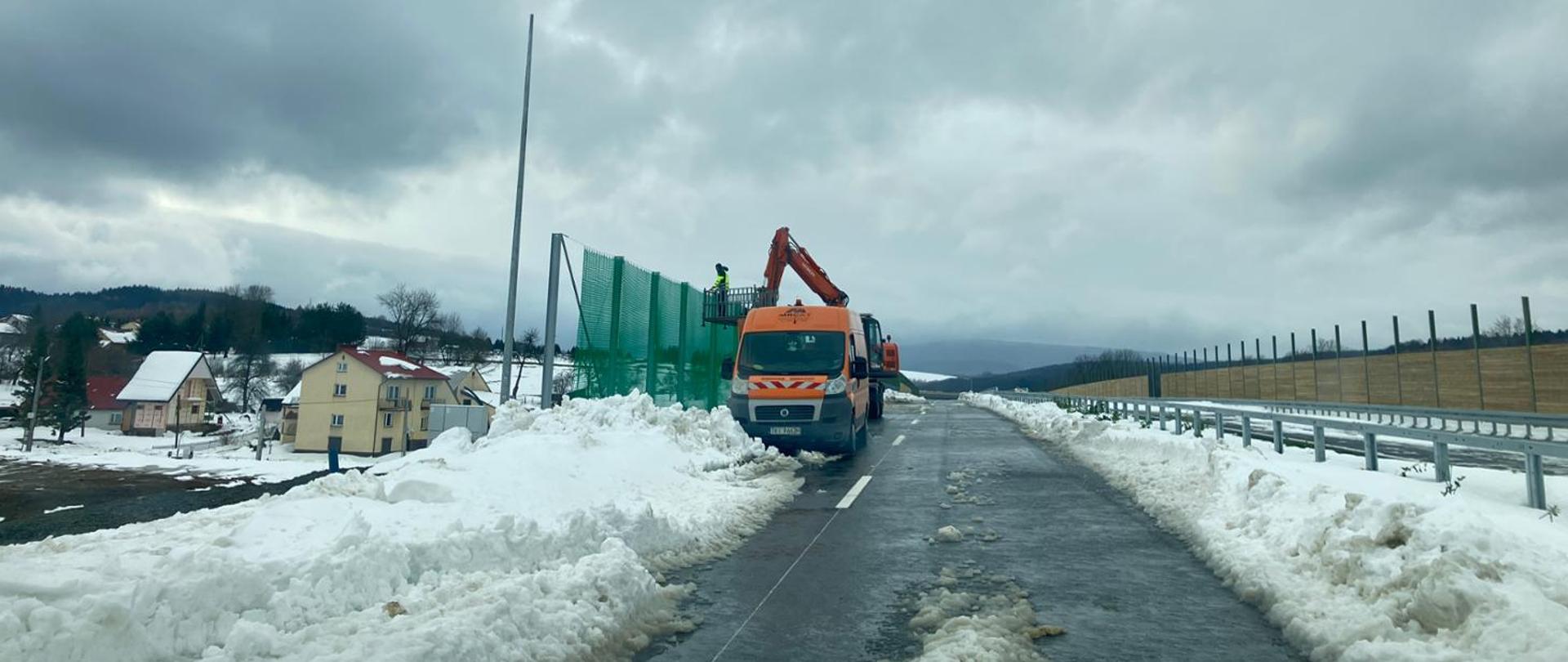 Droga w zimowej scenerii, częściowo odśnieżona, z mokrym śniegiem na nawierzchni. Po obu stronach wysokie zaspy. Na środku stoi pomarańczowa ciężarówka z wysięgnikiem, obok montowane są zielone ekrany akustyczne. W tle domy z ośnieżonymi dachami i pagórkowaty teren. Niebo całkowicie zachmurzone.