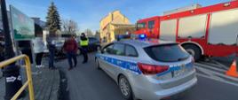 Kolizja w Miejskiej Górce. Droga krajowa nr 36.Na pierwszym planie radiowóz policyjny i wóz strażacki. W głębi funkcjonariusz policji i osoby cywilne. Za nimi pojazdy uczestniczące w kolizji. W tle budynki i drzewa.