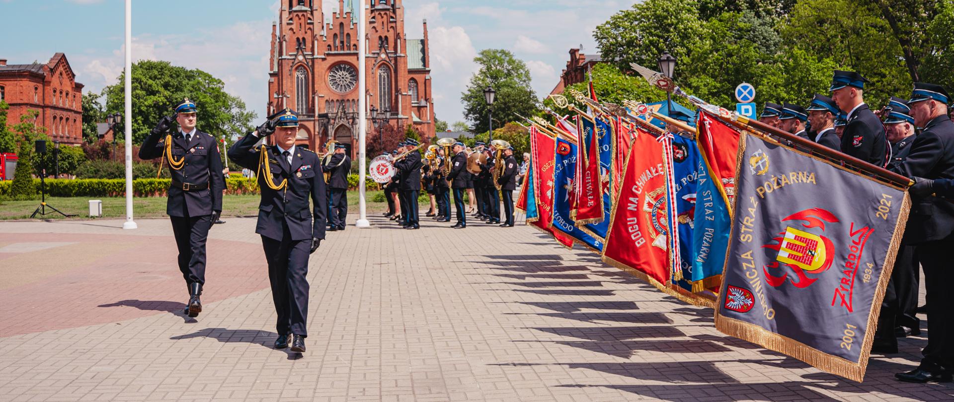 Na zdjęciu widać uroczysty przemarsz strażackich pocztów sztandarowych odbywający się na placu Jana Pawła II. Uczestnicy ubrani w galowe mundury maszerują w uporządkowanym szyku, niosąc czerwone i granatowe sztandary jednostek Państwowej Straży Pożarnej. W tle znajduje się wysoki kościół z charakterystycznymi wieżami oraz drzewa otaczające plac. Pogoda jest słoneczna, na niebie widoczne są jasne chmury, a cała scena ma podniosły i oficjalny charakter związany z obchodami strażackiego święta.