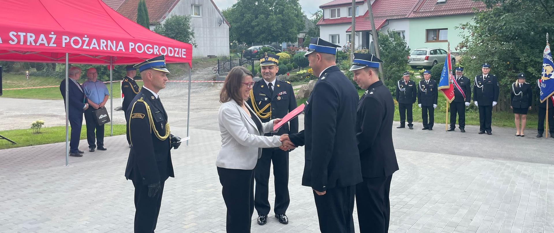 Moment przekazania dokumentów nowego samochodu druhom z Pogrodzia przez panią Burmistrz Tolkmicka. Towarzyszy jej Warmińsko-Mazurski Komendant Wojewódzki oraz Komendant Miejski z Elbląga. Z lewej strony widoczny czerwony namiot, w tle zgromadzeni goście i strażacy.
