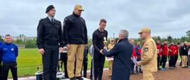 Na zdjęciu przedstawiona jest dekoracja trzech najlepszych drużyn zawodów Mistrzostw Województwa Świętokrzyskiego w Sporcie Pożarniczym. Widoczni są kierownicy drużyn stojący na podjum. Kierownicy drużyn ubrani są w służbowe umundurowanie. Jednemu z nich stojącemu na trzecim miejscu wręczany jest puchar. Puchar wręcza Wojewoda Świętokrzyski wraz Świętokrzyskim Komendantem Wojewódzkim PSP w Kielcach. W dalszym tle widać część zawodników startujących drużyn w zawodach.