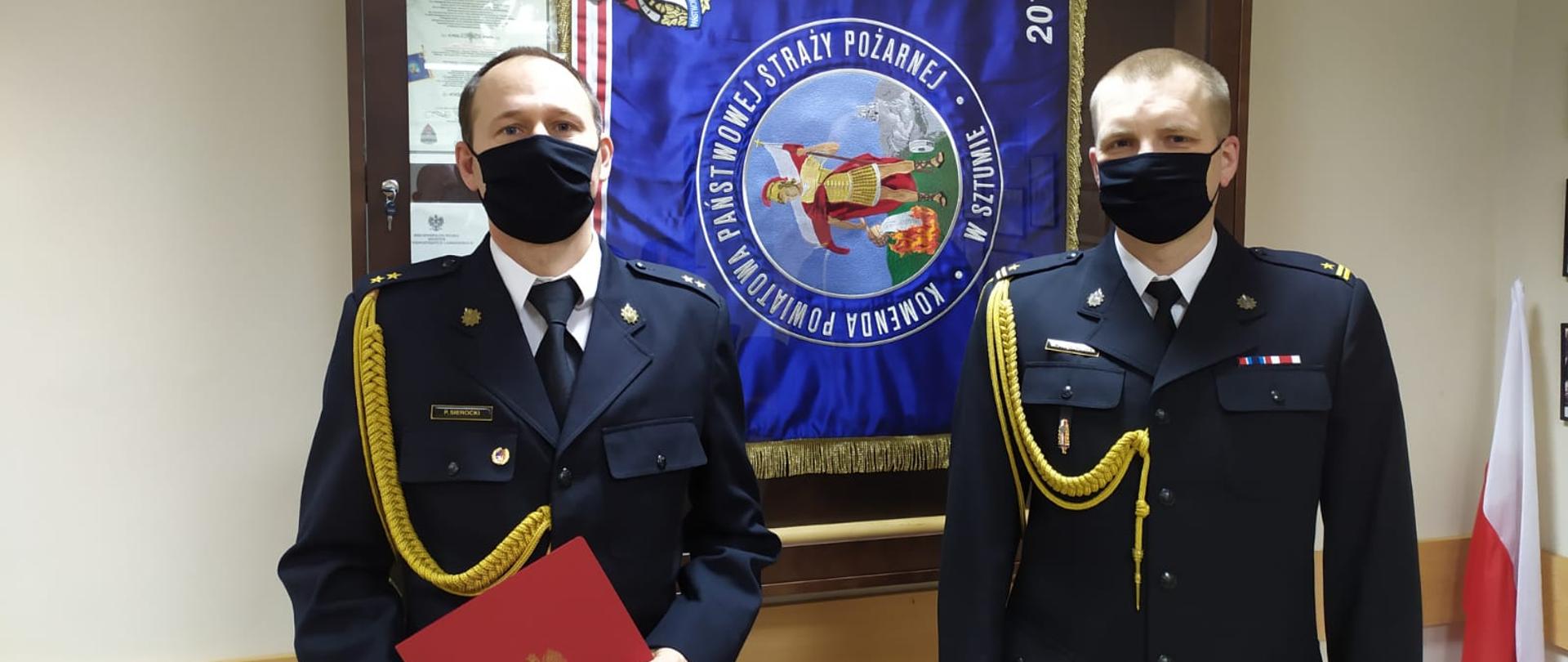 Na zdjęciu stoi dwóch mężczyzn w mundurach galowych, na twarzach maja czarne maseczki ochronne. Mężczyzna po lewej stronie trzyma w rękach przed sobą akt nadania stopnia służbowego. Po prawej stronie zdjęcia stoi zastępca komendanta powiatowego PSP z rękoma opuszczonymi wzdłuż ciała. Obu mężczyzn widać do pasa, a w tle wisi w gablocie sztandar komendy.