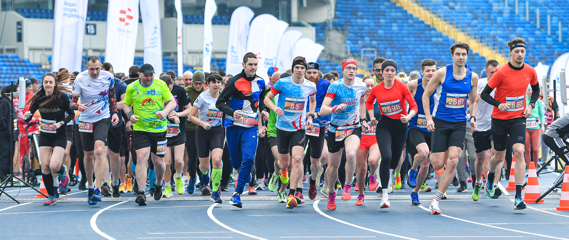 Grupa ludzi rusza do biegu. W tle trybuny i windery.