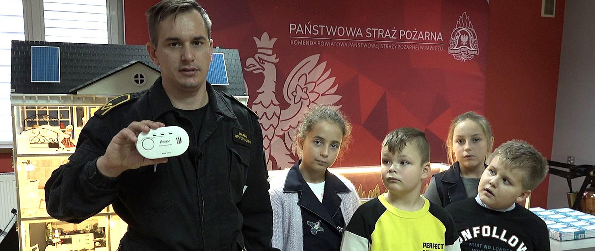 Konkurs "Czujka na straży Twojego bezpieczeństwa". Na pierwszym planie strażak, trzymający w prawej ręce białą czujkę czadu. Obok niego stoi czworo dzieci - dwie dziewczynki i dwóch chłopców. W tle mobilny symulator zagrożeń oraz ścianka reklamowa.
