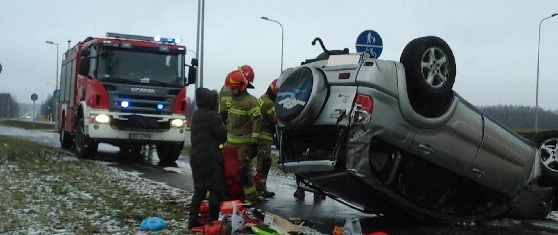 Samochód osobowy leżący na dachu na poboczu jezdni. Przy samochodzie pracujący strażacy. W tle samochód ratowniczo-gaśniczy oraz radiowóz policyjny.
