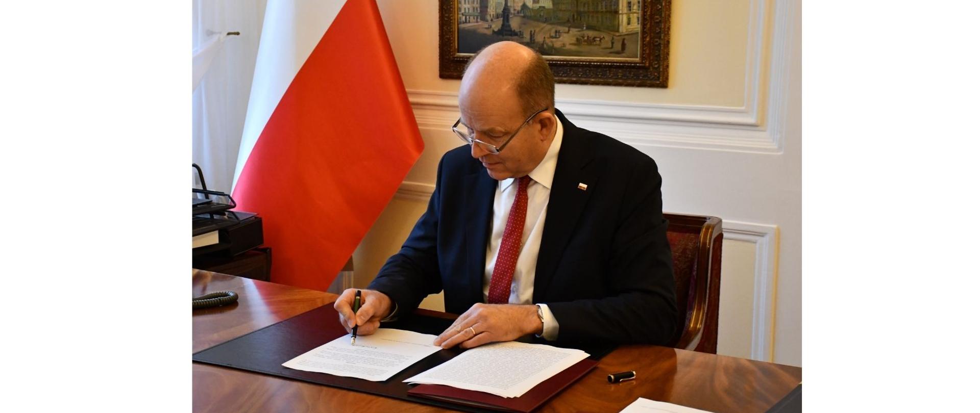 Na pierwszym planie zdjęcia znajduje się elegancko ubrany mężczyzna. Mężczyzna ten siedzi przed biurkiem, podpisując decyzję o pozwoleniu na użytkowanie tunelu drogowego w Warszawie. W tle znajduje się między innymi biało-czerwona flaga oraz wiszący przy ścianie obraz przedstawiający Plac Zamkowy w Warszawie - z Zamkiem Królewskim i Kolumną Zygmunta. 