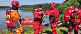 Zdjęcia wykonane jest na brzegu jeziora i przedstawia strażaków, którzy ubrani są w ubrania do pracy w wodzie i kamizelki ratunkowe. Na głowach mają kaski i czapki termiczne. Jeden ze strażaków pomaga drugiemu strażakowi się ubierać. W tle zdjęcia w wodzie widać łódkę strażacką oraz stojące na brzegu samochód strażacki z przyczepką do łodzi. W tle zdjęcia widać brzeg jeziora i drzewa. 