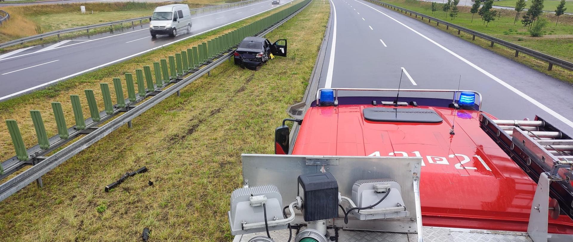 Zdjęcie zrobione z dachu samochodu pożarniczego. Na zdjęciu widać samochód osobowy poza drogą w pozycji na kołach, który stoi na pasie zieleni bezpośrednio przy barierach energochłonnych